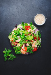 Fresh shrimp salad with cherry tomatoes, arugula, lamb lettuce, cucumber, red onion and sesame seeds on black table background. Top view