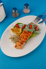 Grilled salmon steak. A piece of red fish on a white plate and a blue background on the table. Fish food with sauce, tomatoes, lemon. Restaurant menu. Grill fried