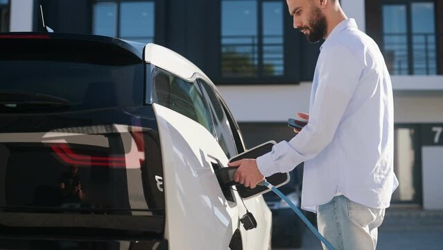 Putting charging wire into the car and using smartphone while waiting. Stylish young man in white clothes is outdoors.