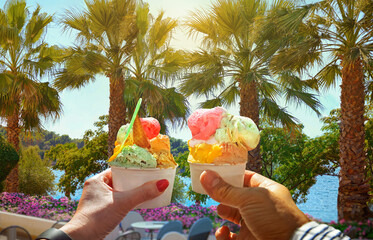 Couple with beautiful bright  sweet  ice-cream with different flavors  in the hands. Background of   view of the see with coconut palm trees in Porec ,Croatia.Traveling concept background - 577304930