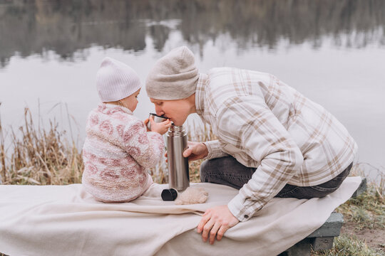 Man and girl are sitting by the river and drinking from thermos