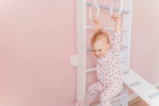 A Caucasian girl in soft pink pajamas does sport at gymnastic rings