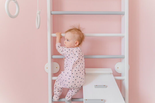 A cute toddler girl in soft pink pajamas does gymnastic on wall bars