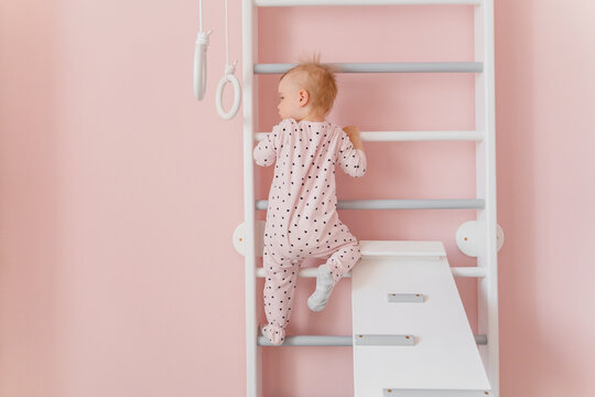 A cute toddler girl in soft pink pajamas does gymnastic on wall bars