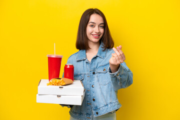 Young Ukrainian woman holding fast food isolated on yellow background making money gesture