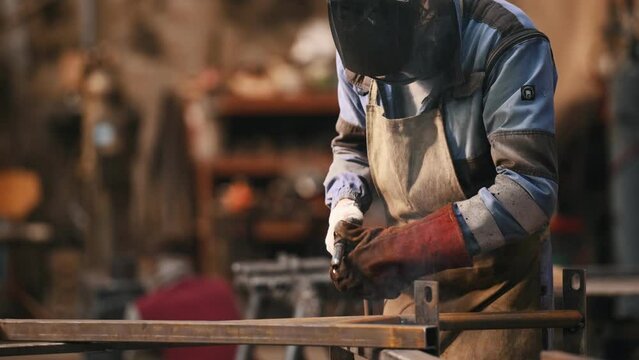 Another man is walking behind. Close up view. Factory worker in protective mask is welding the iron.
