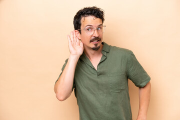 Young caucasian man isolated on beige background listening to something by putting hand on the ear