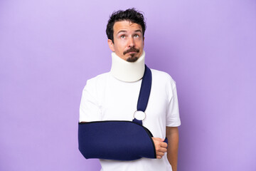 Young caucasian man wearing a sling and neck brace isolated on purple background and looking up