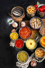 Canned vegetables, beans, fish and fruits in tin cans on black background. Food stocks.