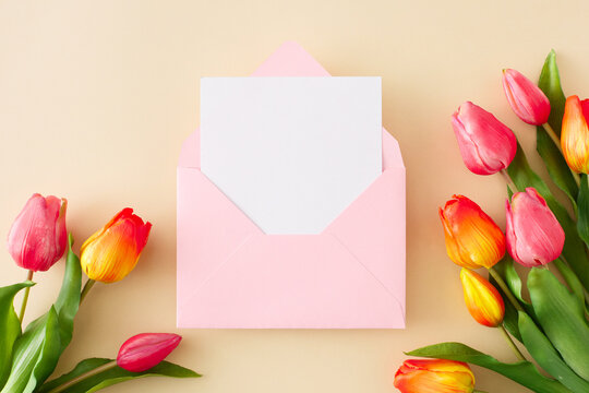 Hello spring concept. Flat lay photo of open pink envelope with card red tulips flowers on isolated beige background. Holiday card idea