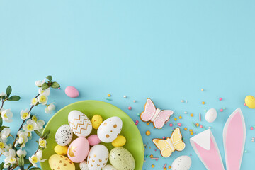 Easter decoration concept. Top view composition of green plate with colorful eggs easter bunny ears gingerbread sprinkles and cherry blossom branch on isolated pastel blue background with empty space