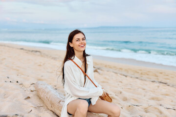 Fototapeta na wymiar Happy tanned woman smiling with teeth in white swimsuit shirt and denim shorts sits on the sand by the ocean with wet hair after swimming, sunset light and pink clouds in Bali