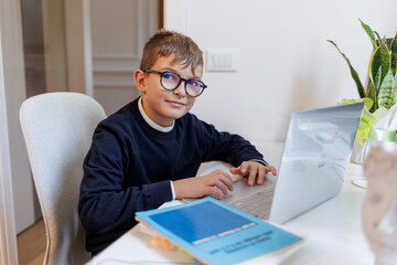 Bambino biondo con gli occhiali , vestito in blu e seduto davanti a una scrivania mentre lavora al computer