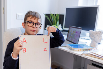 Bambino biondo con gli occhiali , vestito in blu e seduto davanti a una scrivania , mostra una...