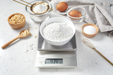 Flour in white bowl measuring on digital scale with cake or bakery ingredients and utensil on marble kitchen table