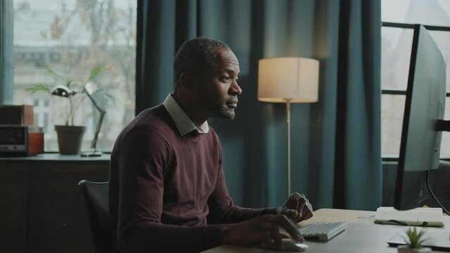 Footage of thoughtful African American man working in front of computer at home. Employee typing on keyboard, looking at monitor in cozy living room. Indoors