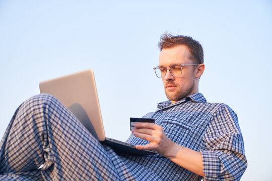Online shopping: caucasian bearded male in eyeglasses making shopping outdoor using laptop and credit card wearing nightwear. Clear blue sky at background. High quality image