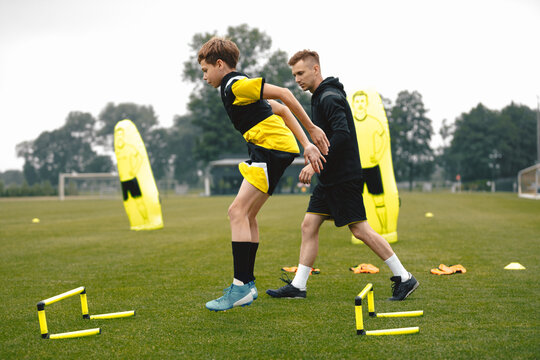 Teenage Boy at Training Drill With Young Soccer Coach. Youth Football Player Jumping Over Row of Hurdles at Sports Practice Field. Sports Strength and Agility Training For Footballers