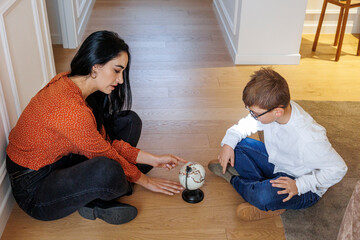 Ragazza adulta  con i capelli scuri mostra a un bambino un mappamondo seduti nel pavimento di casa © alex.pin