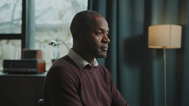 Side view of focused African man wearing comfortable clothes sitting at desktop with computer and looking at screen. Businessman searching information online in peaceful and cozy atmosphere of home