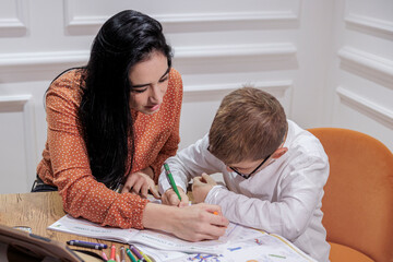 Un educatrice con i capelli scuri aiuta nel fare i compiti un bambino biondo con gli occhiali, all'interno di  in un ambiente elegante.