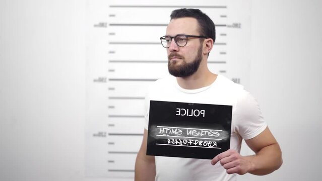 Side profile mugshot of man holding sign while being photographed in police department. Realtime