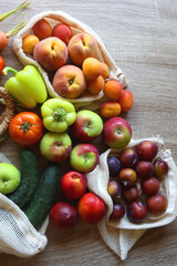 Straw bag and reusable fabric bags filled with various healthy fruit and vegetables. Wooden background, top view.