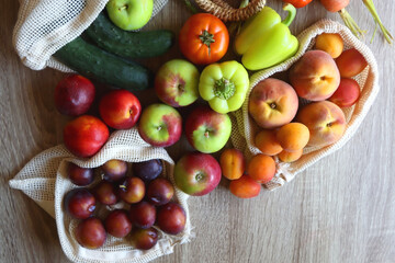Straw bag and reusable fabric bags filled with various healthy fruit and vegetables. Wooden background, top view.