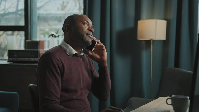 Shot of irritated African man sitting in front of computer and having difficult conversation on smartphone. Businessman gesturing actively, talking to business partner. Indoors. Modern interior of