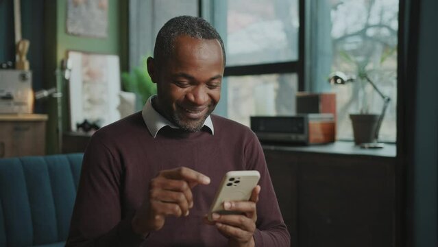 Joyful pleasant-looking African American man using smartphone, checking messages, chatting with friends on social media during lunch break. Smiling male freelancer working from home