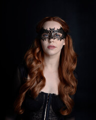 Naklejka premium Masked close up portrait of beautiful woman with long red hair wearing sheer corset top, and black bat wing mask. Isolated on dark studio background with. Moody silhouette lighting.