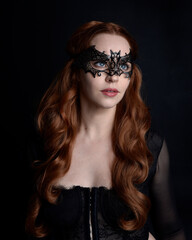 Naklejka premium Masked close up portrait of beautiful woman with long red hair wearing sheer corset top, and black bat wing mask. Isolated on dark studio background with. Moody silhouette lighting.