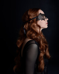 Fototapeta premium Masked close up portrait of beautiful woman with long red hair wearing sheer corset top, and black bat wing mask. Isolated on dark studio background with. Moody silhouette lighting.