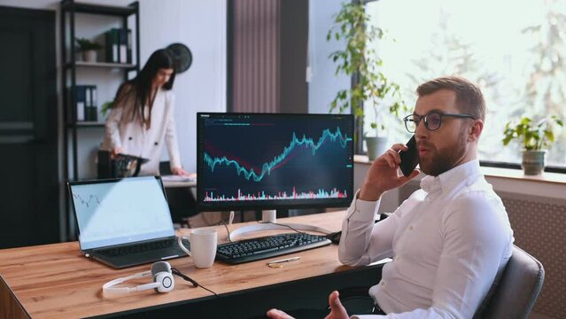 Stock broker is sitting by computer with graphs and talking by phone. Woman is at background.