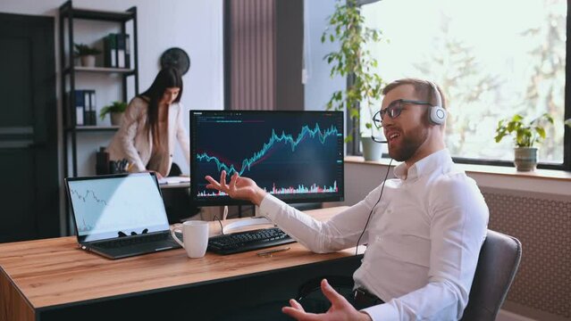 Stock broker is sitting by computer with graphs and talking in headphones with microphone. Woman is at background.