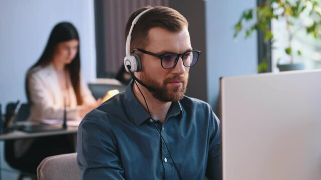 Man in headphones with microphone is working by a computer in the office. Woman is behind.
