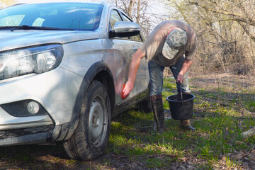 A man in a cap washes the car with a sponge and water from a bucket