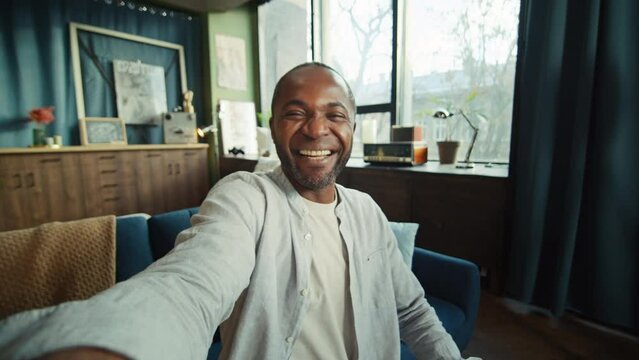 Portrait of happy emotional African American man talking through videocall, waving and looking at camera of smartphone. Excited man talking to friend while sitting on sofa in living room. Daytime
