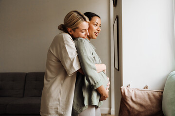 Pregnant lesbian couple hugging and looking through window while standing together at home