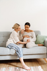 Pregnant lesbian couple holding hands and looking at each other while sitting on couch