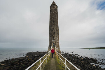 Chaine Memorial Tower, "The Pencil" Larne, Northern Ireland, UK, with beach, ocean and cloudy sky with bi racial hiker woman in red jacket and white beanie hat - Powered by Adobe