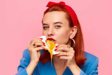 Close-up portrait of young redhead woman eating fried eggs against pink studio background. Liquid yolk. Food pop art photography. Breakfast. Complementary colors. Copy space for ad, text