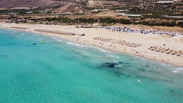 Aerial view descending to Falassarna beach with tourist sunbathing on sandy Greek island and swimming in transparent clear ocean water