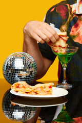 Close-up image of male hand putting pizza slice into cocktail against yellow studio background. Party time. Food pop art photography, creativity. Complementary colors. Copy space for ad, text