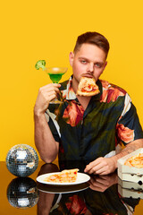 Food pop art. Young emotionless man in stylish shirt sitting at table, drinking cocktail and eating pizza against yellow studio background. Party time. Complementary colors. Copy space for ad, text