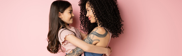 Side view of smiling kid hugging tattooed mom on pink background, banner.