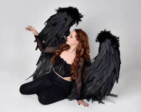 Full length portrait of beautiful woman with long red hair wearing sheer corset top, leather pants & large black angel feather wings.  Sitting pose with gestural hands reaching out, kneeling on floor.