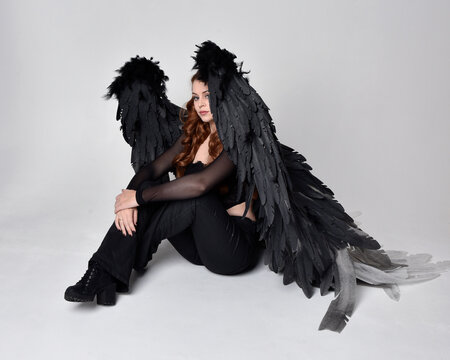 Full length portrait of beautiful woman with long red hair wearing sheer corset top, leather pants & large black angel feather wings.  Sitting pose with gestural hands reaching out, kneeling on floor.