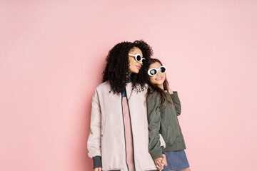 Cheerful preteen kid in sunglasses holding hand of stylish mom on pink background.
