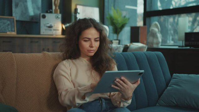 Horizontal footage of attractive young elegant woman with cheerful smile looking directly at camera. Cute smiling girl touching screen of gadget, reading messages indoor. Relax and comfort